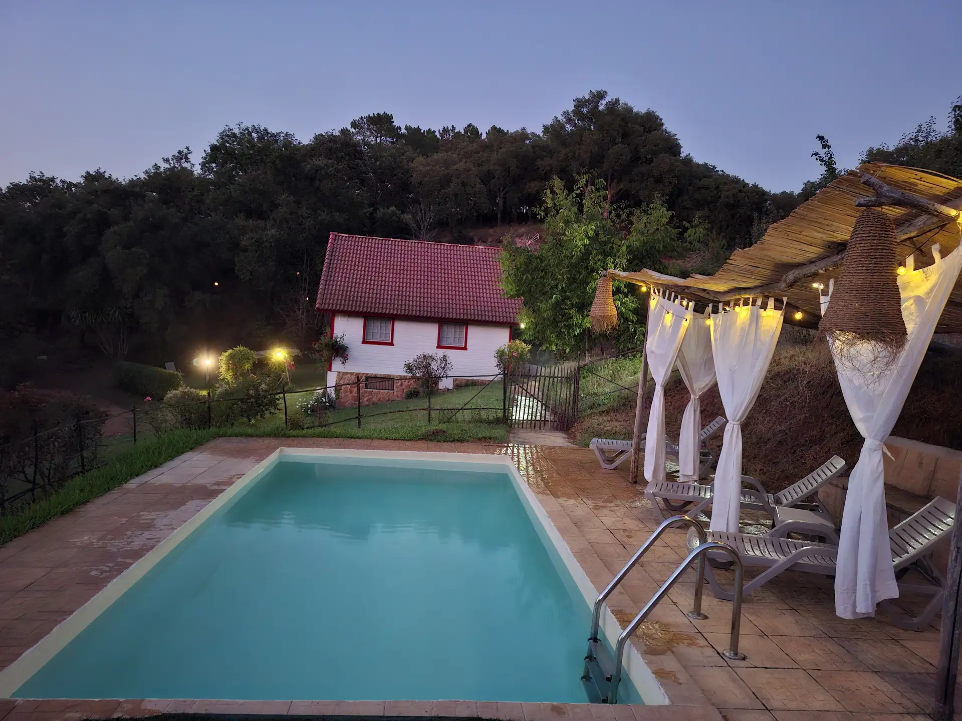 Vista de la piscina con el sombrajo, y la casa al fondo al anochecer